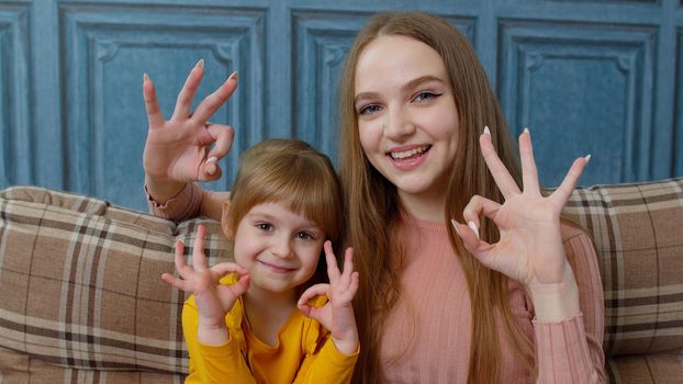 Portrait of happy little child kid daughter with young mother, hugging, embracing, showing ok gesture sign while sitting on sofa at home. Girl enjoying sweet moment with smiling mom in living room