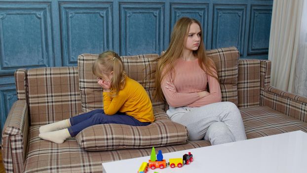 Unhappy stressed woman and angry little child sister ignoring each other sitting on couch after quarrel. Upset girl sibling avoid talk sitting turned back on sofa at home having argument with family