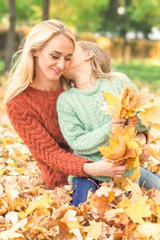 Happy young caucasian mother and little daughter holding autumn yellow leaves sitting and kissing at the park