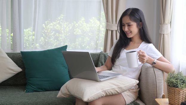 Attractive asian woman sitting in living room and checking email on her laptop.