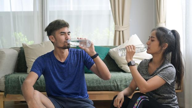Young couple in sports clothes is drinking water, resting on the floor after workout.