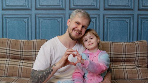Father and little child daughter kid in pajamas sit on couch in room smiling, looking at camera, joyfully making sign of shape heart near chest together. Girl with young man. Bonding family generation