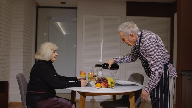 Senior retired couple having fun drinking wine and eating healty meal during romantic supper sitting in the kitchen celebrating their anniversary. Mature man and woman wife on active elderly festivity
