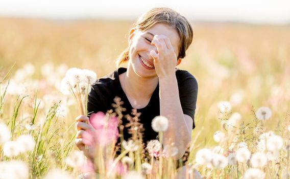 Pretty girl teenager sitting in field with dandelions and flowers, looking at camera and smiling outdoors. Beautiful young female person at nature in sunny day portrait