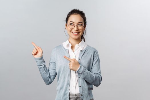 Education, studying and online classes concept. Intrigued, excited happy asian woman in glasses peeking left, smiling and pointing upper corner at advertisement or chart, grey background.