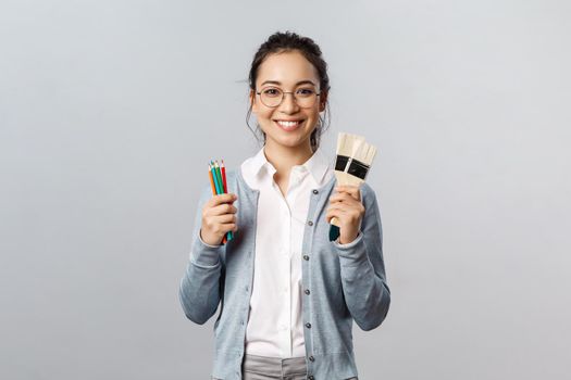 Hobbies, creativity and art concept. Cheerful young asian woman in glasses spend leisure with fun, holding colored pencils and two painting brushes, smiling upbeat, drawing artwork.