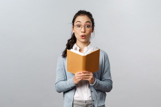 Education, teachers, university and schools concept. Amused and excited asian female tutor reading good homework of student in notebook, fold lips and look camera wondered, grey background.