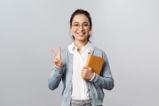 Education, teachers, university and schools concept. Cheerful asian female brunette, student or tutor holding planner notebooks and show peace sign, smiling optimistic, grey background.
