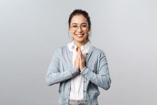 Emotions, people and lifestyle concept. Friendly cute asian woman in glasses, hold hands in pray or namaste gesture, smiling cheerful, greeting person politely, standing grey background.