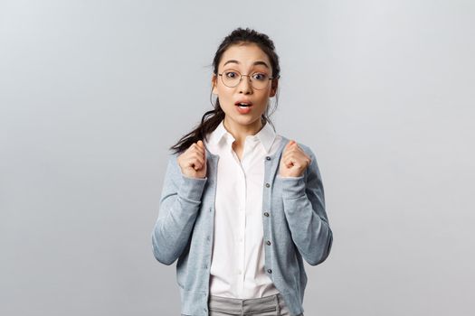 Portrait of excited and tempting cute asian girl in glasses, awaiting with thrill and interest for new season of favorite tv show coming up, looking ancitipation and amusement, stand grey background.
