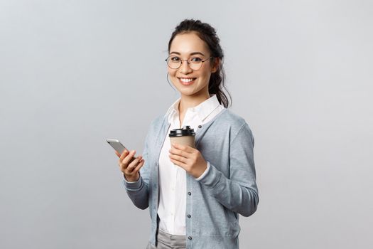 Office lifestyle, business and people concept. Cheerful attractive asian girl in glasses, holding take-away coffee and smartphone, look pleased at camera with smile, contact person videocall chat.