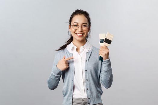 Creativity, repairs and overhaul concept. Cheerful smiling asian woman pointing at painting brushes as renovating her living room, picking paint for walls during repairment works, grey background.