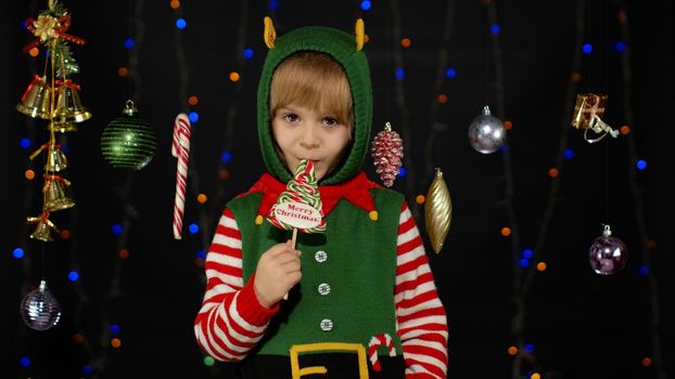 Blonde kid girl in Christmas elf Santa Claus helper costume licking candy lollipop sweets isolated on black background with garland. Child enjoying caramel sweets. People New Year holidays celebration