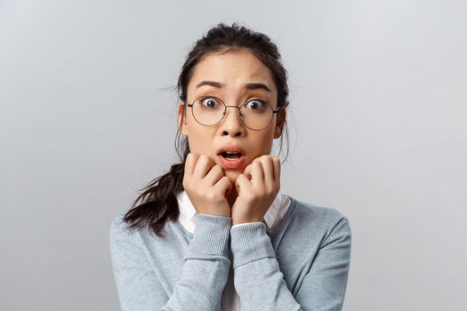 Close-up portrait alarmed, afraid young asian woman heart terrifying bad news, hold hands pressed to face, gasping and open mouth, say oh my gosh as staring camera scared, grey background.