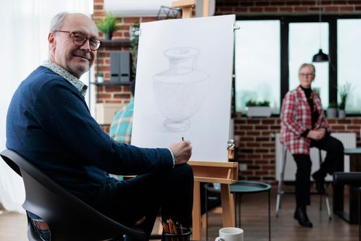 Portrait of smiling senior artist standing in front on white canvas drawing artistic vase using sketching technique during art lesson in creativity studio. Elderly painter havnig creative hobby