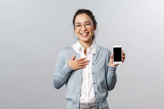 Technology, online and mobile lifestyle concept. Happy boastful asian woman showing pictures or her profile on app smartphone display, holding phone laughing and smiling camera pleased.