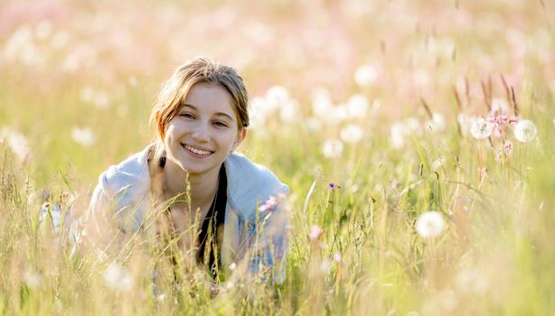 Pretty girl teenager sitting in field with dandelions and smiling outdoors. Beautiful young female person at nature with flowers