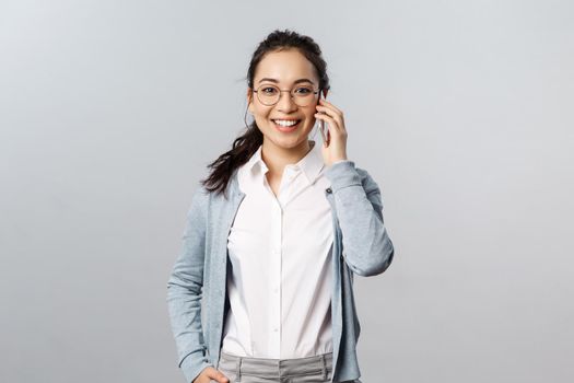 Office lifestyle, business and people concept. Friendly attractive asian female in glasses, calling friend during quarantine, stay in touch connected with family, use mobile phone and smile.