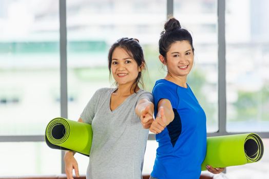 Asian adult and young woman smiling holding a yoga mat after yoga and exercise and show thumb up sign. Portrait of happy beautiful female hold yoga mat indoor studio, sport healthy workout concept