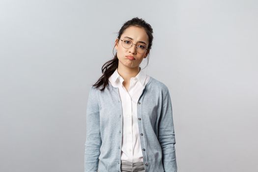 Portrait of sighing displeased and gloomy young asian girl in glasses, pouting and looking disappointed camera, express regret or feel uneasy over failure at work, grey background.