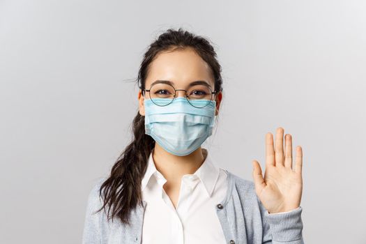 Covid19, virus, health and medicine concept. Portrait of friendly young girl in medical face mask saying hi, hello waving hand and grinning with eyes, keep safe during pandemia, quarantine.