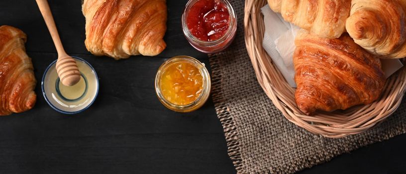 Delicious, fresh croissants in wicker basket with honey and jams on black wooden table for breakfast.