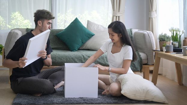 Happy young couple sitting in floor and unpacking cardboard boxes at new home. Relocation house renovation, removals concept.