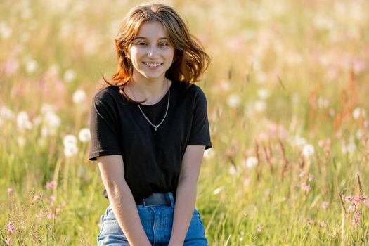 Pretty girl teenager sitting in field with dandelions and smiling outdoors. Beautiful young female person at nature in sunny day portrait