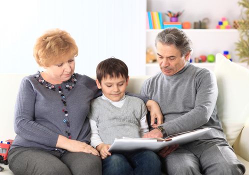 Cute boy sitting in the lap of their grandparents and looking happily together at a photo album