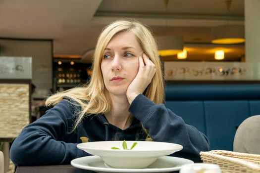 Young lonely sad pensive blonde woman in a cafe with a plate of food. Selective focus.