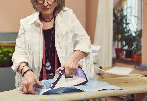 Mature white-haired Caucasian woman seamstress, tailor ironing blue shirt with steaming on the iron board before trying in on a mannequin in fashion design atelier