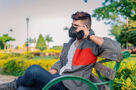 Man sitting on a bench calling on the phone, young man with a mask calling on the cell phone, man with a mask calling on the phone in a park