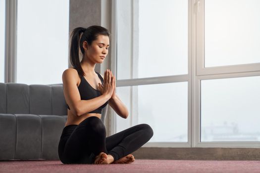 Side view of brunette girl in black leggings sitting in position, meditating with namaste hands at home. Attractive lady with closed eyes and folded palms practicing yoga along. Concept of meditation.