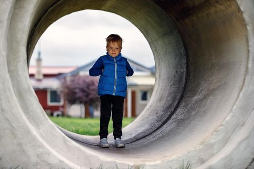 A cute kid boy, wearing a blue vest, stands in a large concrete tunnel. Children's games in the yard.