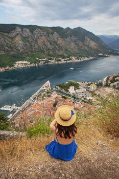 A girl in a blue dress and a straw hat sits against the backdrop of the Bay of Kotor on a beautiful summer day in Montenegro. A very beautiful fjord