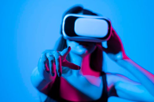 Cheerful girl with hands up wearing the virtual reality goggles and playing a games in neon light. Future technology concept. Metaverse