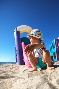 A little boy in a hat play with sand in a beach, and looks to sea