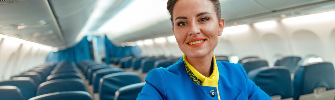 Joyful female flight attendant or air hostess looking at camera and smiling while placing hands on passenger seat