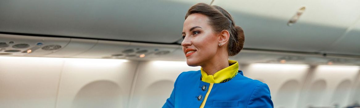 Cheerful woman stewardess in air hostess uniform standing near passenger seat in airplane salon