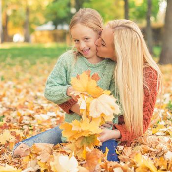 Happy young caucasian mother and little daughter holding autumn yellow leaves sitting and kissing at the park