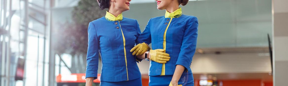 Two cheerful women stewardesses in aviation air hostess uniform looking at each other and smiling while holding travel bags