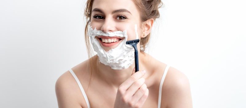 Beautiful young caucasian woman shaving her face by razor on white background. Pretty woman with shaving foam on her face