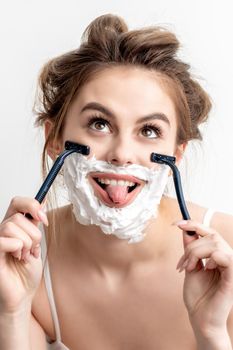 Beautiful young caucasian woman shaving her face by two razors with tongue out on white background.