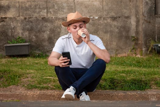 Young man sitting on sidewalk checking phone, man drinking coffee while checking his phone