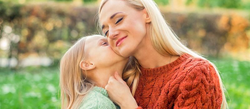 Cute daughter kisses her young mother while sitting at the park