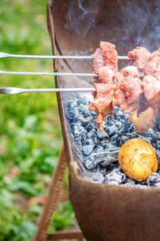 Hands of man prepares barbecue meat with potatoes on skewer by grill on fire outdoors. Concept of lifestyle rustic food preparation