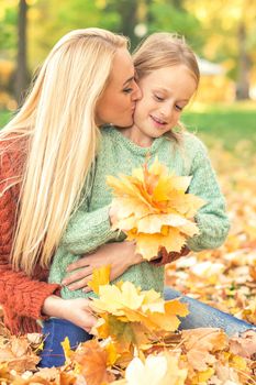 Happy young caucasian mother and little daughter holding autumn yellow leaves sitting and kissing at the park