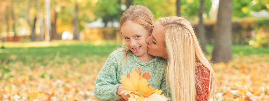 Happy young caucasian mother and little daughter holding autumn yellow leaves sitting and kissing at the park