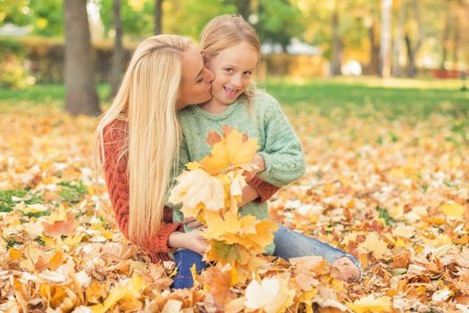 Happy young caucasian mother and little daughter holding autumn yellow leaves sitting and kissing at the park