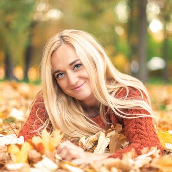 Beautiful young blond hair caucasian woman lies down on leaves at the autumn park